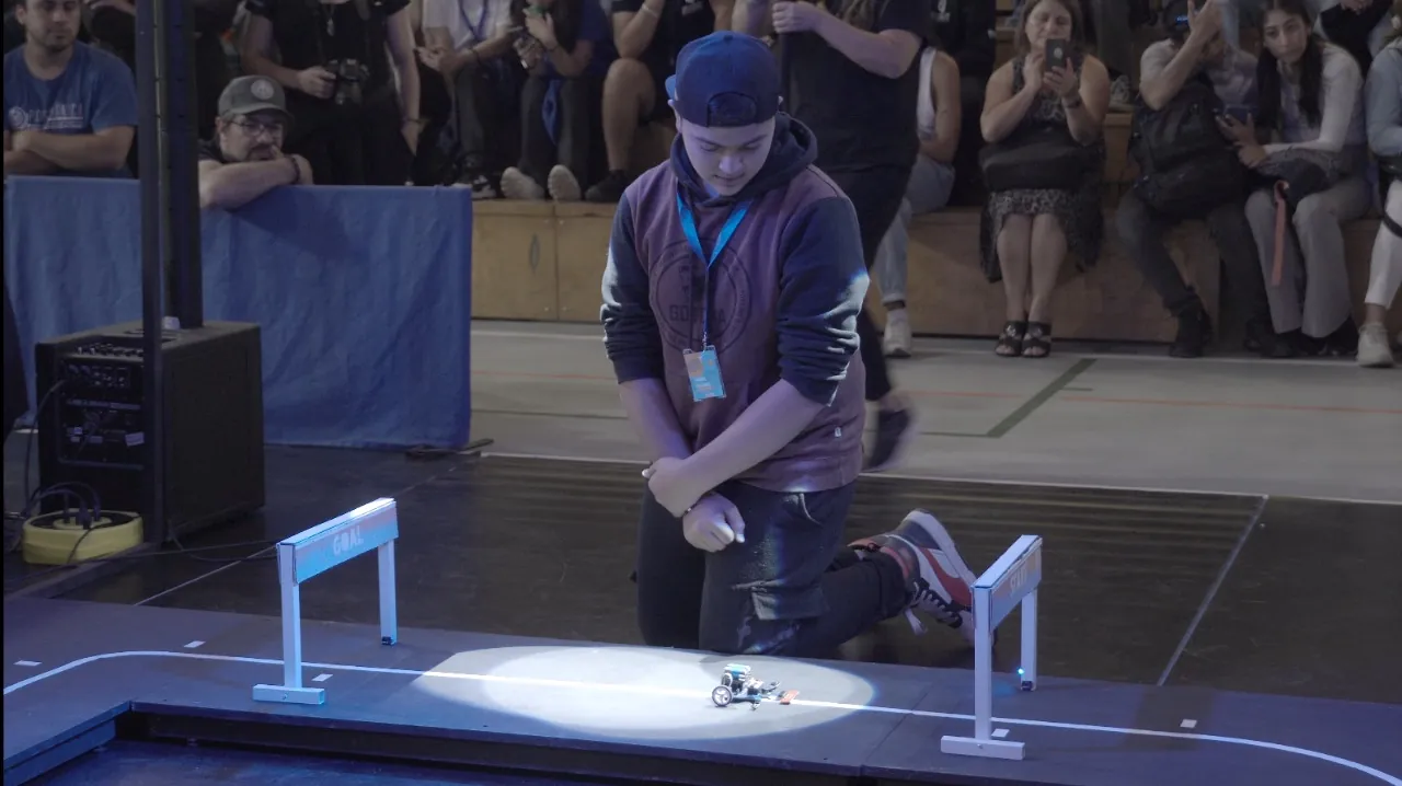 Un competidor suelta su robot en la partida de la pista, bajo un foco, mientras el público lo graba con sus teléfonos